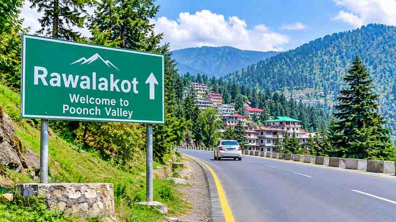 Scenic mountain road leading to Rawalakot, Azad Kashmir, Pakistan, with lush green pine forests, beautiful hills, and a welcoming roadside sign.
